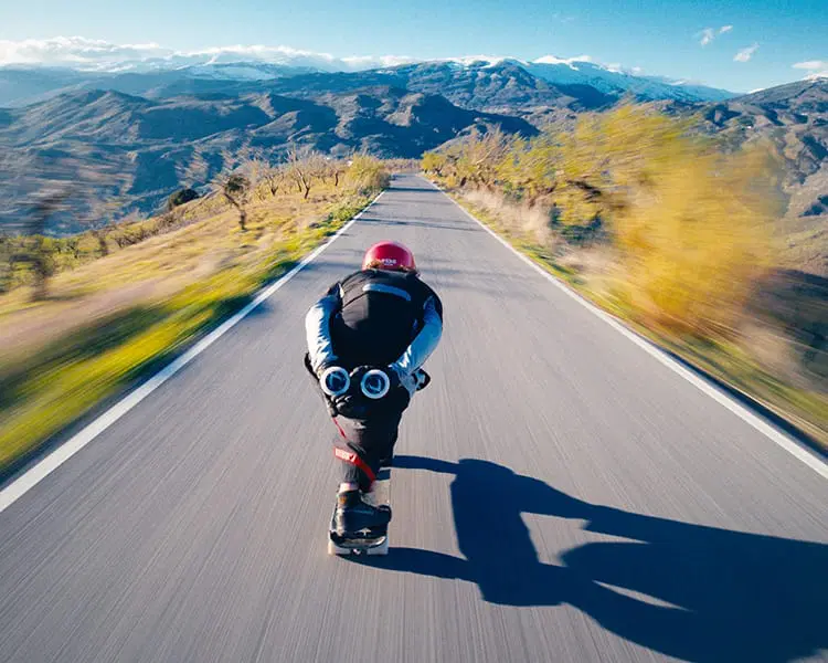 A person wearing a helmet and protective gear skateboards down a winding mountain road at high speed, participating in the Blue Programme, with blurred scenery indicating motion.