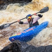 A kayaker wearing a helmet and life jacket maneuvers through rough whitewater rapids in a blue Red Programme kayak branded with Red Bull and Werner logos.