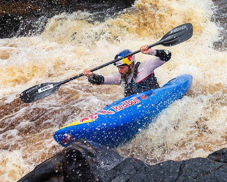 A kayaker wearing a helmet and life jacket maneuvers through rough whitewater rapids in a blue Red Programme kayak branded with Red Bull and Werner logos.