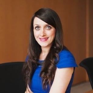 Woman with long brown hair wearing a blue dress sits indoors against a plain brown wall, smiling at the camera.