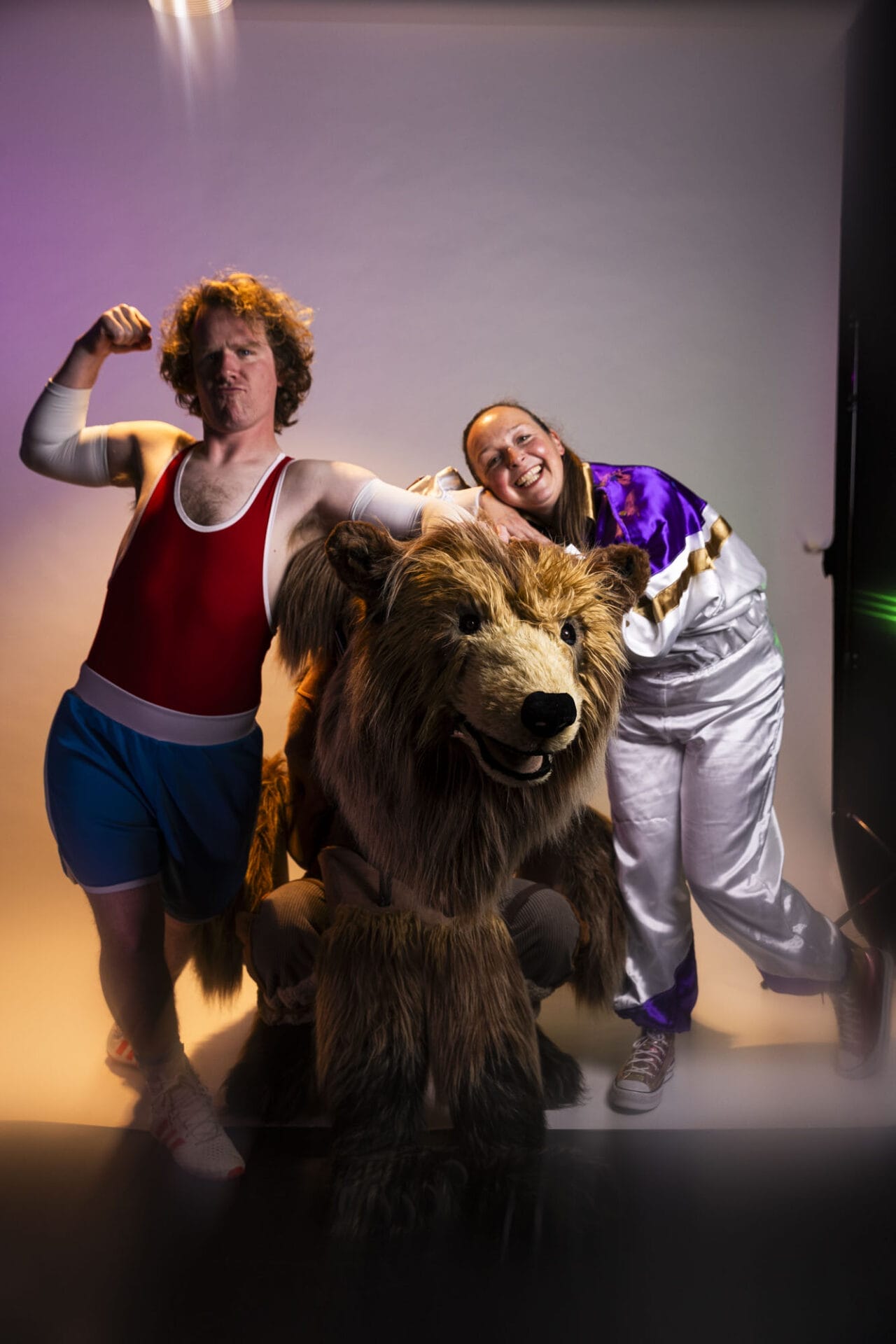 Two people pose with Hercules the Bear; one flexes in athletic wear, the other smiles in a tracksuit, all against a lit studio backdrop.