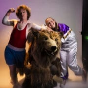 Two people pose with Hercules the Bear; one flexes in athletic wear, the other smiles in a tracksuit, all against a lit studio backdrop.