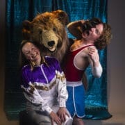 Two people pose with someone in a lion costume. One wears a tracksuit and smiles; the other, dressed in athletic gear, playfully pretends to have their head in the lion’s mouth like Hercules the Bear wrestling his famous foe.