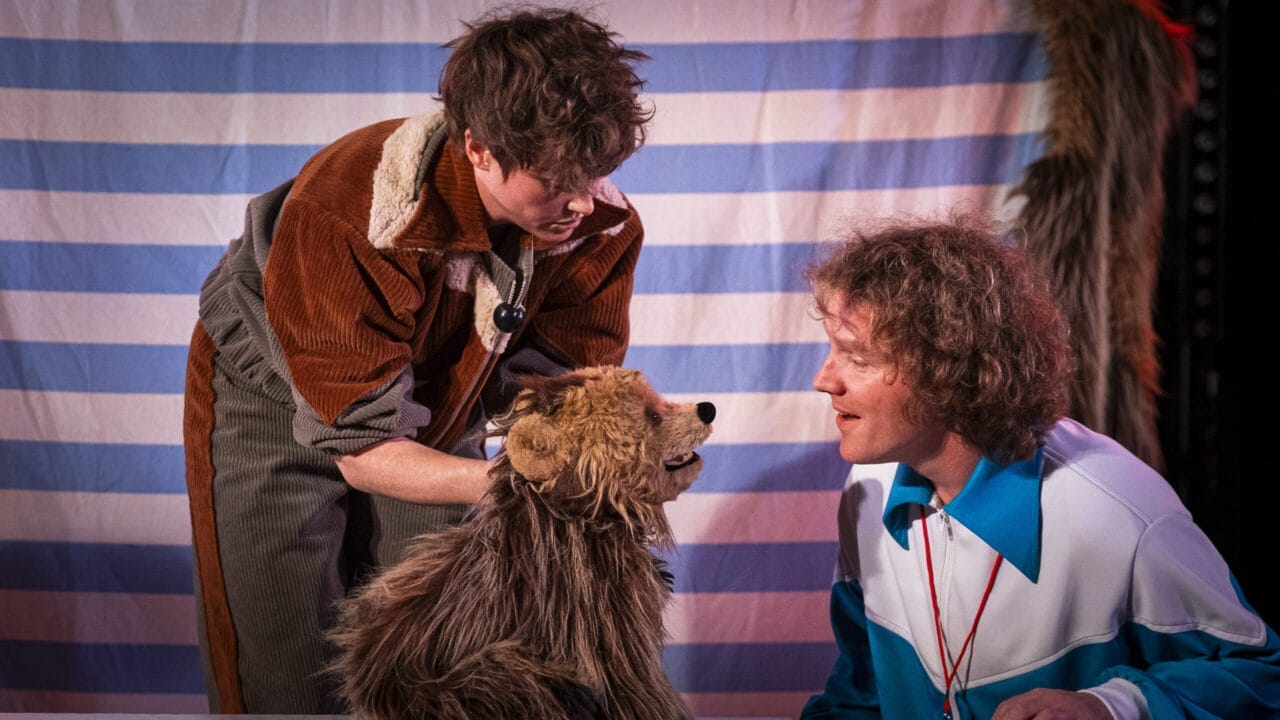 Two people interact with a brown furry puppet that resembles a dog, set against a blue and white striped backdrop, evoking the playful spirit of Hercules the Bear.