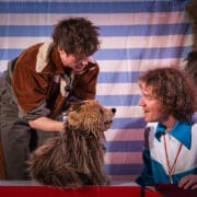 Two people interact with a brown furry puppet that resembles a dog, set against a blue and white striped backdrop, evoking the playful spirit of Hercules the Bear.