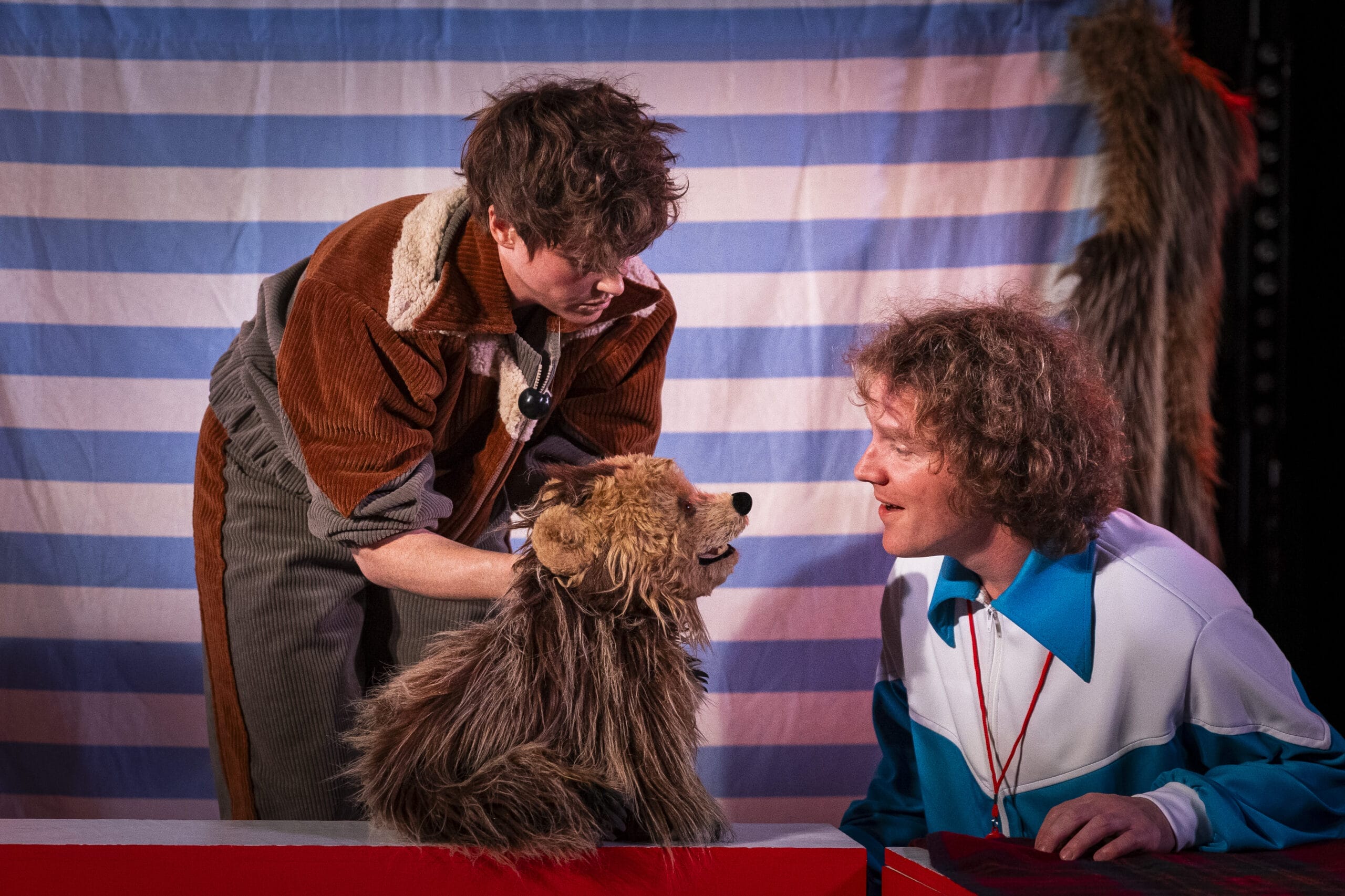 Two people interact with a brown furry puppet that resembles a dog, set against a blue and white striped backdrop, evoking the playful spirit of Hercules the Bear.