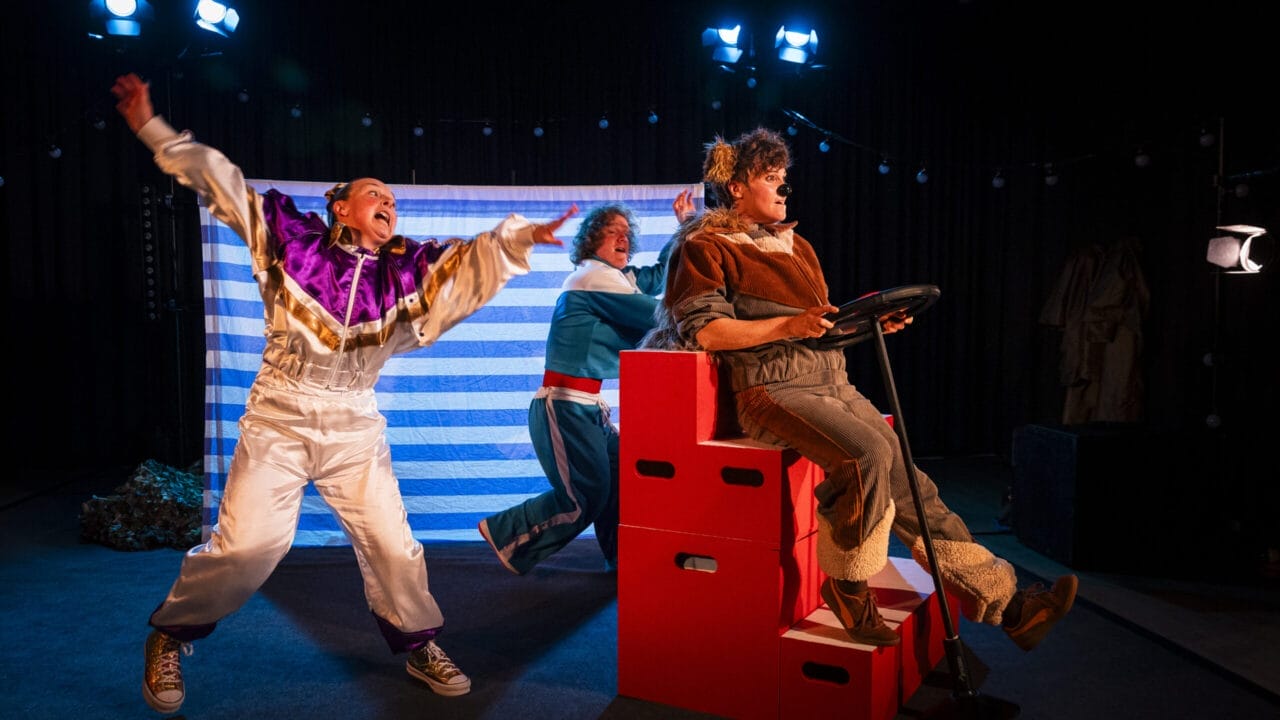 Three actors perform on stage; one wears a clown costume and gestures dramatically, another pushes a red box car like Hercules the Bear, while the third pretends to drive, all beneath bright stage lights.