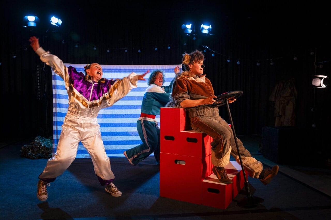 Three actors perform on stage; one wears a clown costume and gestures dramatically, another pushes a red box car like Hercules the Bear, while the third pretends to drive, all beneath bright stage lights.