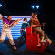 Three actors perform on stage; one wears a clown costume and gestures dramatically, another pushes a red box car like Hercules the Bear, while the third pretends to drive, all beneath bright stage lights.