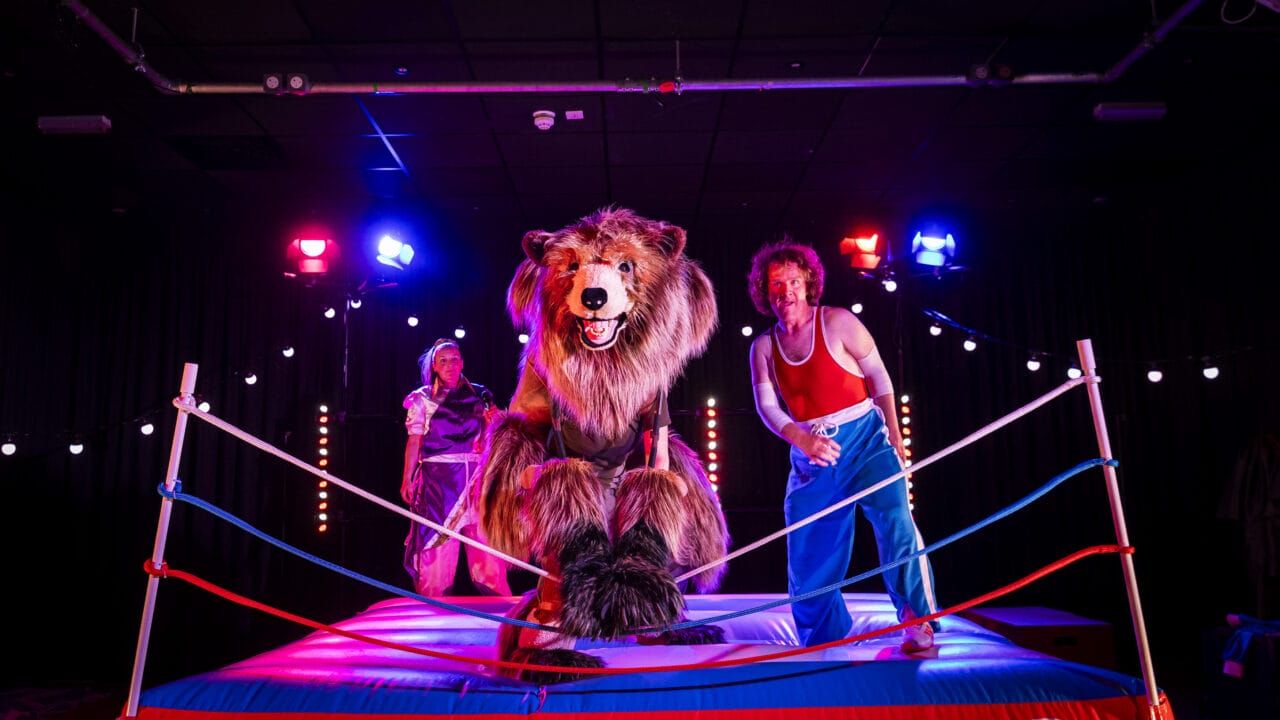 A large bear costume character and two performers stand in a brightly lit wrestling ring with “Hercules” on the apron, surrounded by stage lights.