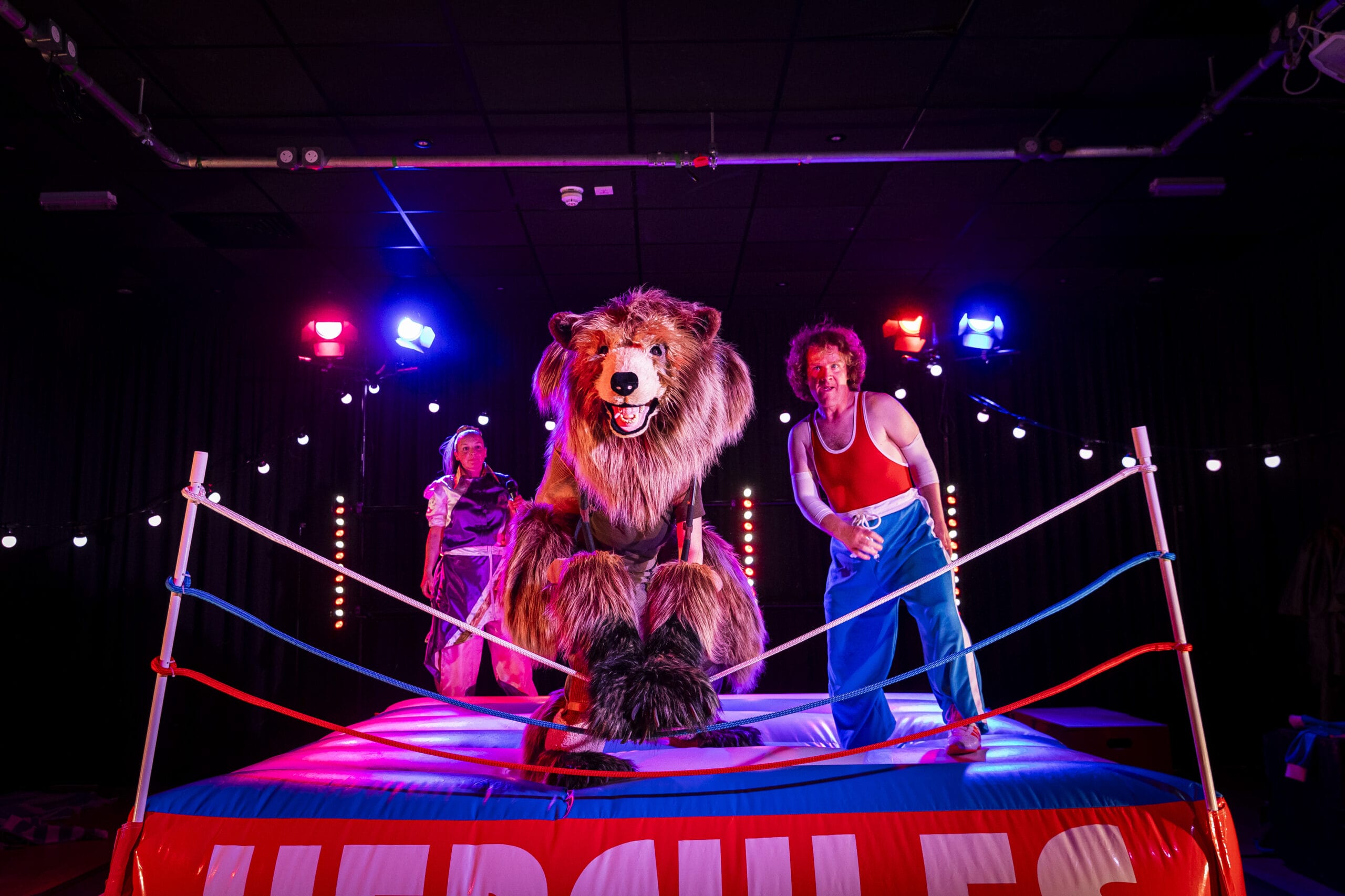 A large bear costume character and two performers stand in a brightly lit wrestling ring with “Hercules” on the apron, surrounded by stage lights.