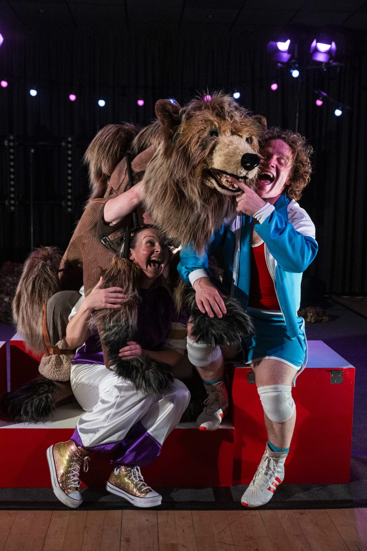 Two performers pose with Hercules the Bear, a large bear puppet, onstage, smiling and playfully interacting with him in a brightly lit theater setting.