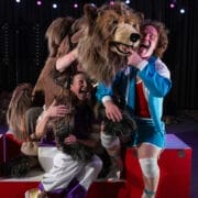 Two performers pose with Hercules the Bear, a large bear puppet, onstage, smiling and playfully interacting with him in a brightly lit theater setting.