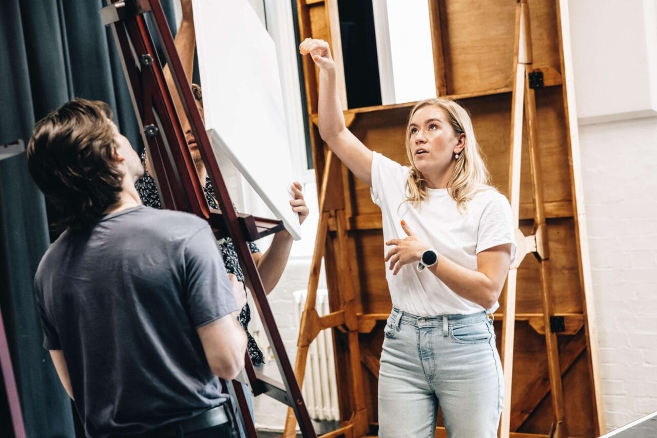 A woman in a white shirt gestures while speaking to a man in a dark shirt, evoking the creative atmosphere of The Talented Mr. Ripley, in a studio setting with easels and art boards in the background.