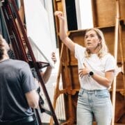 A woman in a white shirt gestures while speaking to a man in a dark shirt, evoking the creative atmosphere of The Talented Mr. Ripley, in a studio setting with easels and art boards in the background.