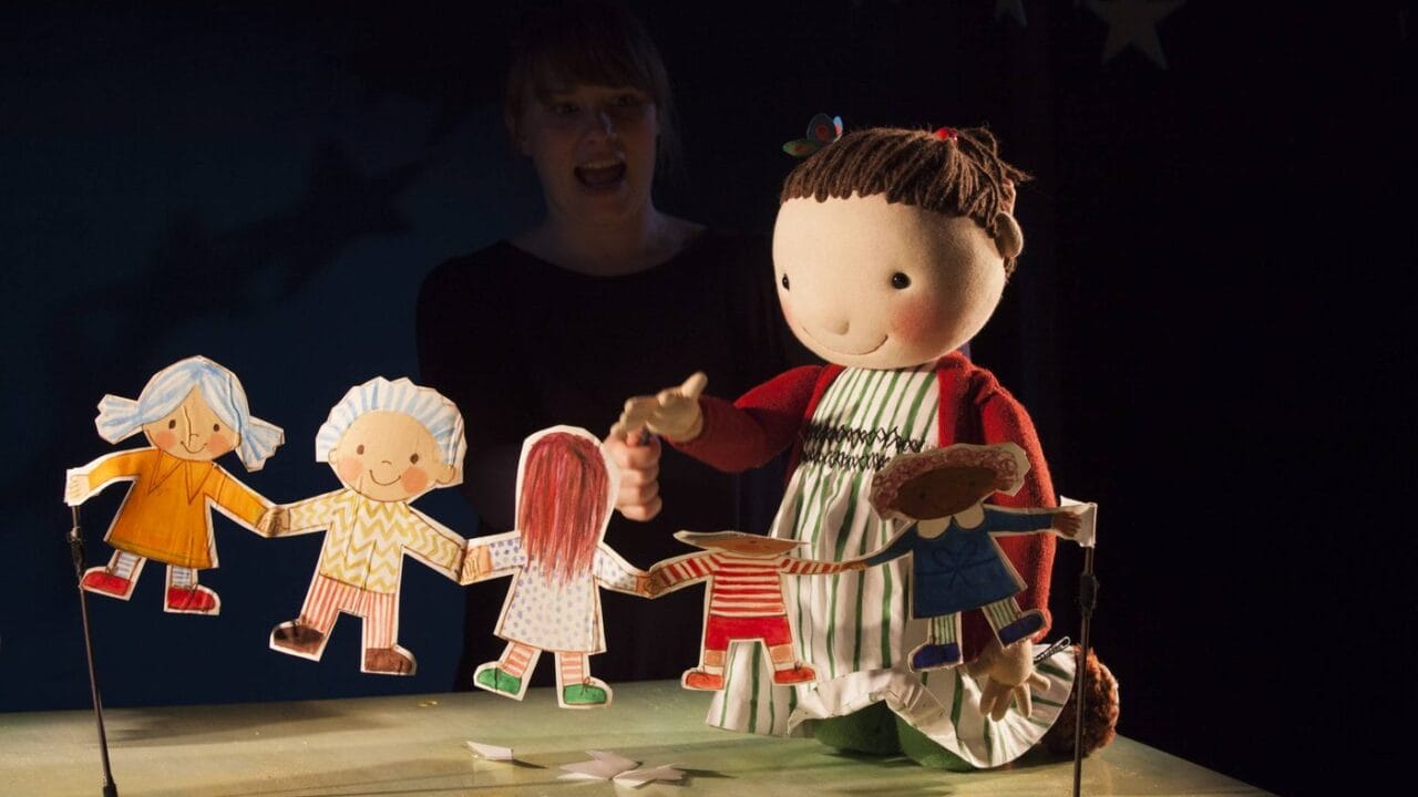 A puppeteer operates a large doll and paper cut-out children on a tabletop stage with a dark, starry background.