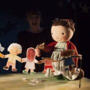 A puppeteer operates a large doll and paper cut-out children on a tabletop stage with a dark, starry background.