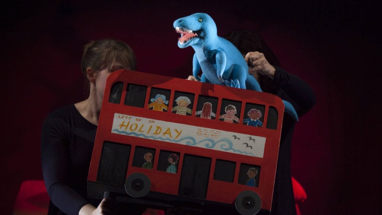 Two people operate a puppet show featuring a red double-decker bus with paper passengers and a blue dinosaur puppet on top.