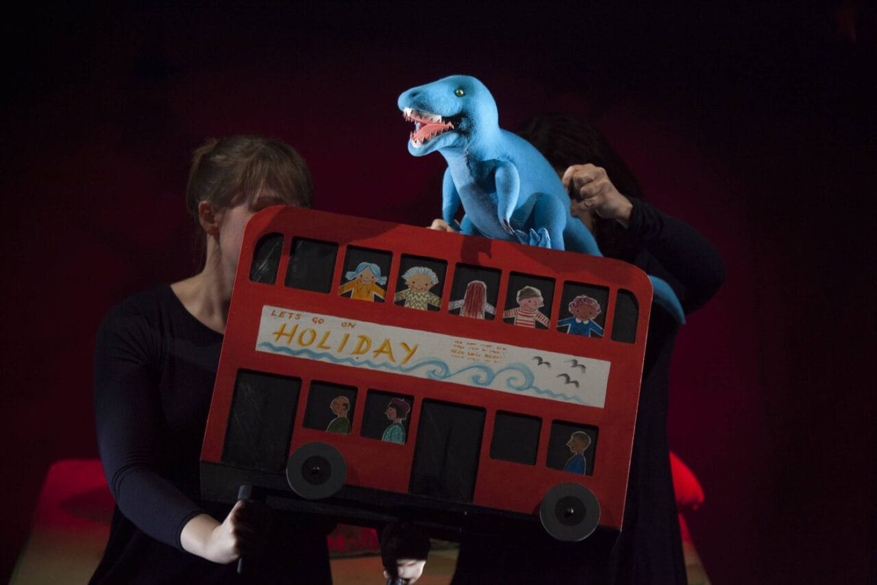 Two people operate a puppet show featuring a red double-decker bus with paper passengers and a blue dinosaur puppet on top.