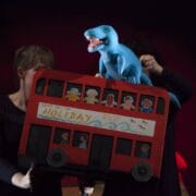 Two people operate a puppet show featuring a red double-decker bus with paper passengers and a blue dinosaur puppet on top.