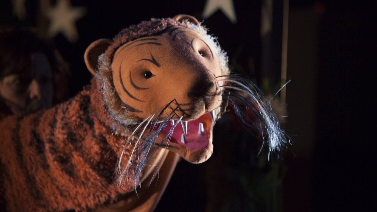 A close-up of a person operating a large, realistic tiger puppet with painted features and visible whiskers, set against a dark background with star decorations.