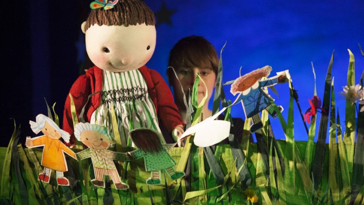 A person operates a large doll puppet dressed in a red sweater and striped dress among paper cut-out figures and tall artificial grass on a stage.