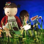 A person operates a large doll puppet dressed in a red sweater and striped dress among paper cut-out figures and tall artificial grass on a stage.