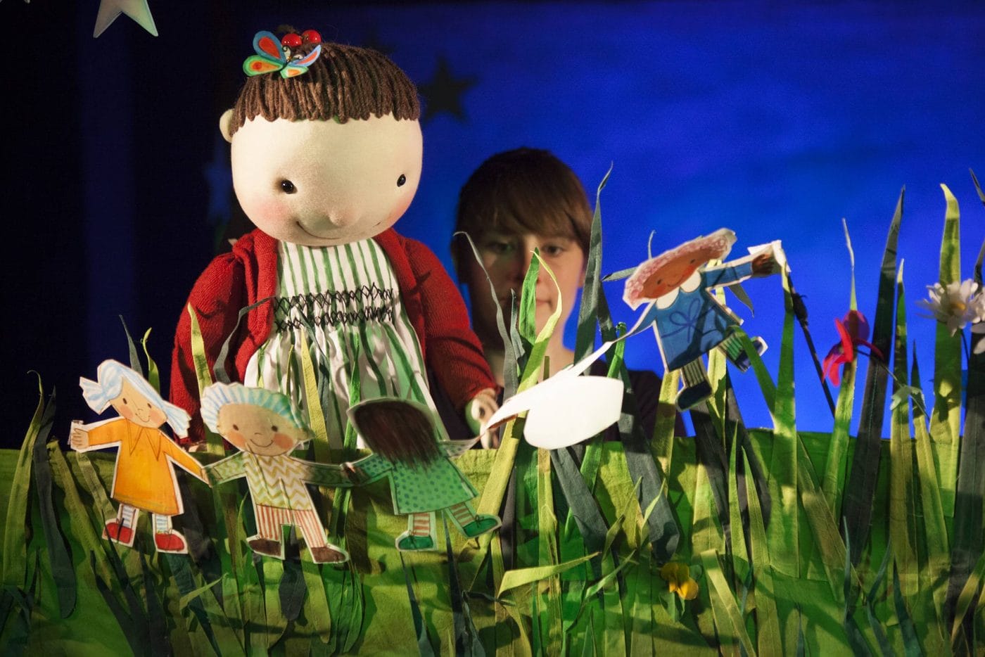 A person operates a large doll puppet dressed in a red sweater and striped dress among paper cut-out figures and tall artificial grass on a stage.