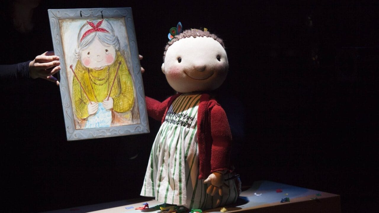 A large puppet of a child in a striped dress and red sweater stands on a table next to a drawing of an older woman held by a person whose face is not visible.