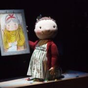 A large puppet of a child in a striped dress and red sweater stands on a table next to a drawing of an older woman held by a person whose face is not visible.