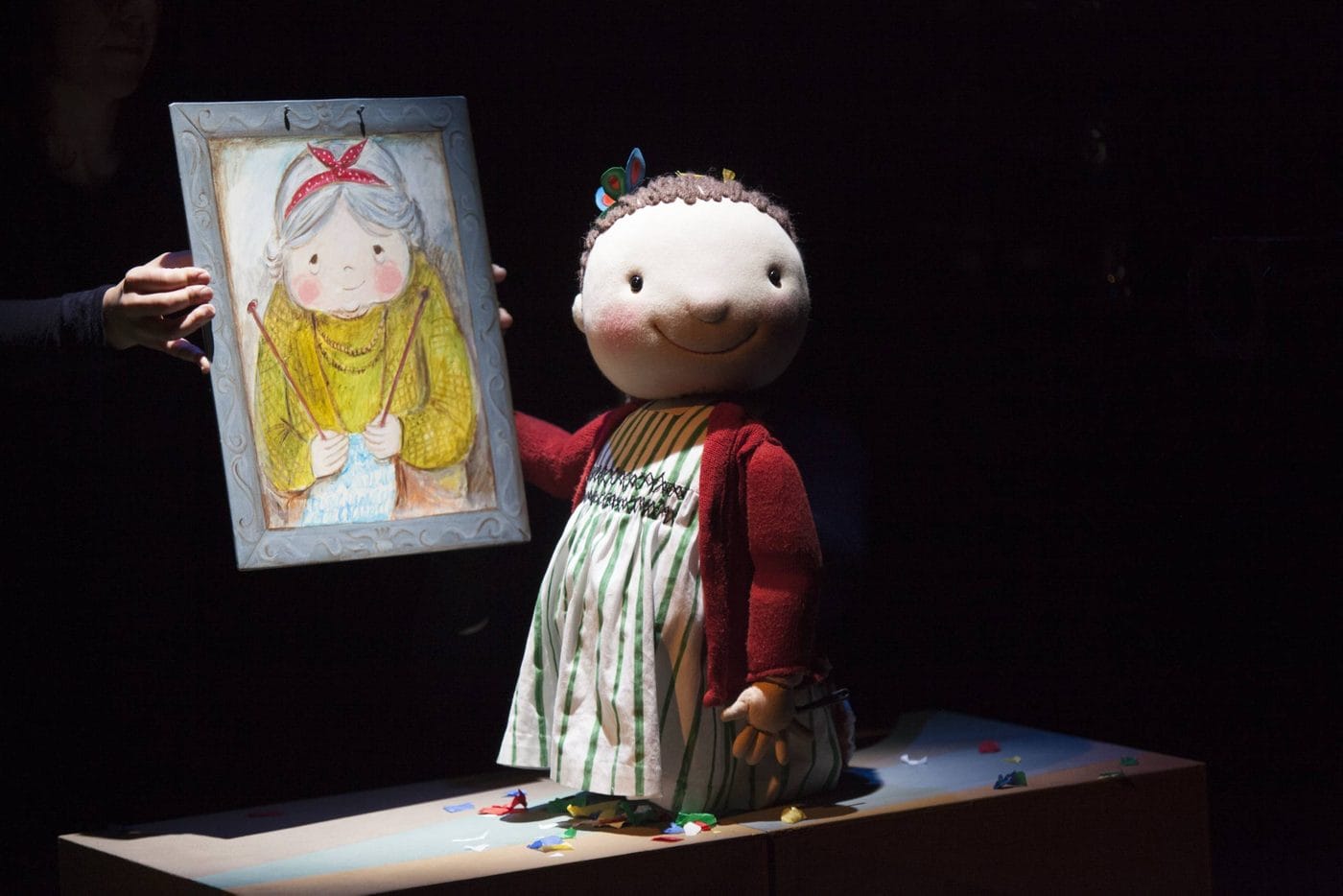 A large puppet of a child in a striped dress and red sweater stands on a table next to a drawing of an older woman held by a person whose face is not visible.