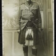Postcard of J.D.Swanston in 1868, wearing a kilt, sporran, knee-high socks, and a cap. Standing indoors holding a cane, with a painted backdrop of flowers behind him.