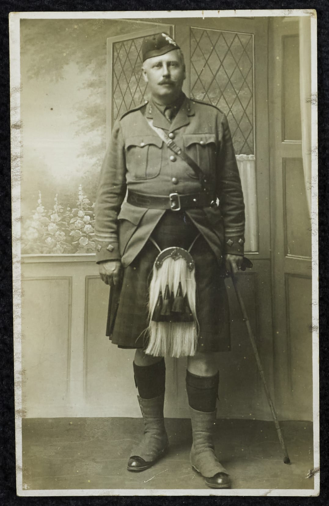 Postcard of J.D.Swanston in 1868, wearing a kilt, sporran, knee-high socks, and a cap. Standing indoors holding a cane, with a painted backdrop of flowers behind him.