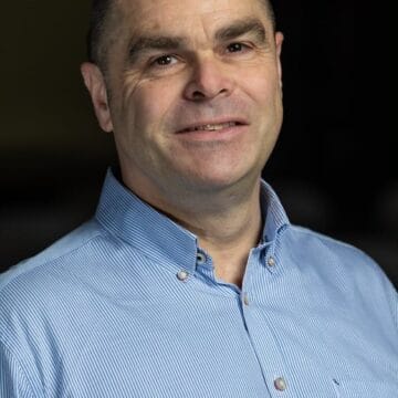 A middle-aged man with short dark hair, wearing a light blue striped button-down shirt, smiling at the camera against a dark background.