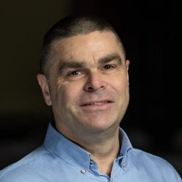 A middle-aged man with short dark hair, wearing a light blue striped button-down shirt, smiling at the camera against a dark background.