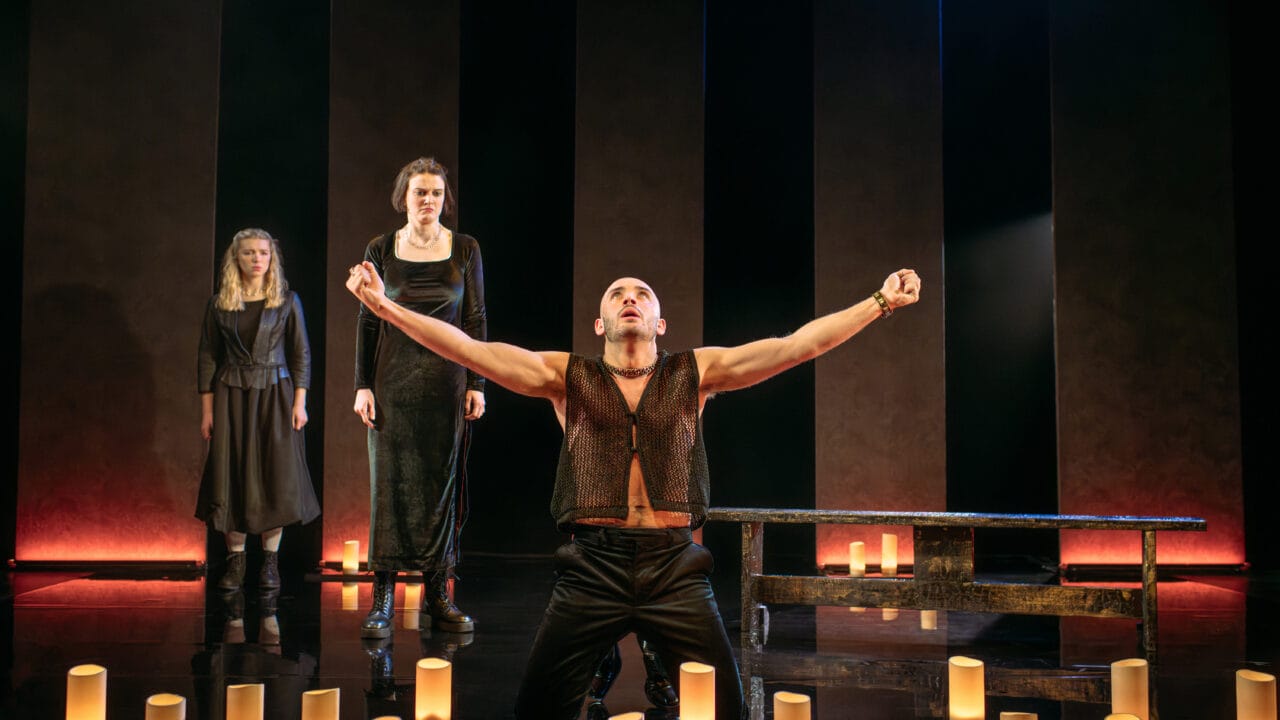 Three actors perform on stage; one man kneels with arms raised and two women stand behind him. The stage is dimly lit with cylindrical lights on the floor.