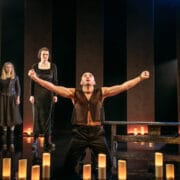Three actors perform on stage; one man kneels with arms raised and two women stand behind him. The stage is dimly lit with cylindrical lights on the floor.