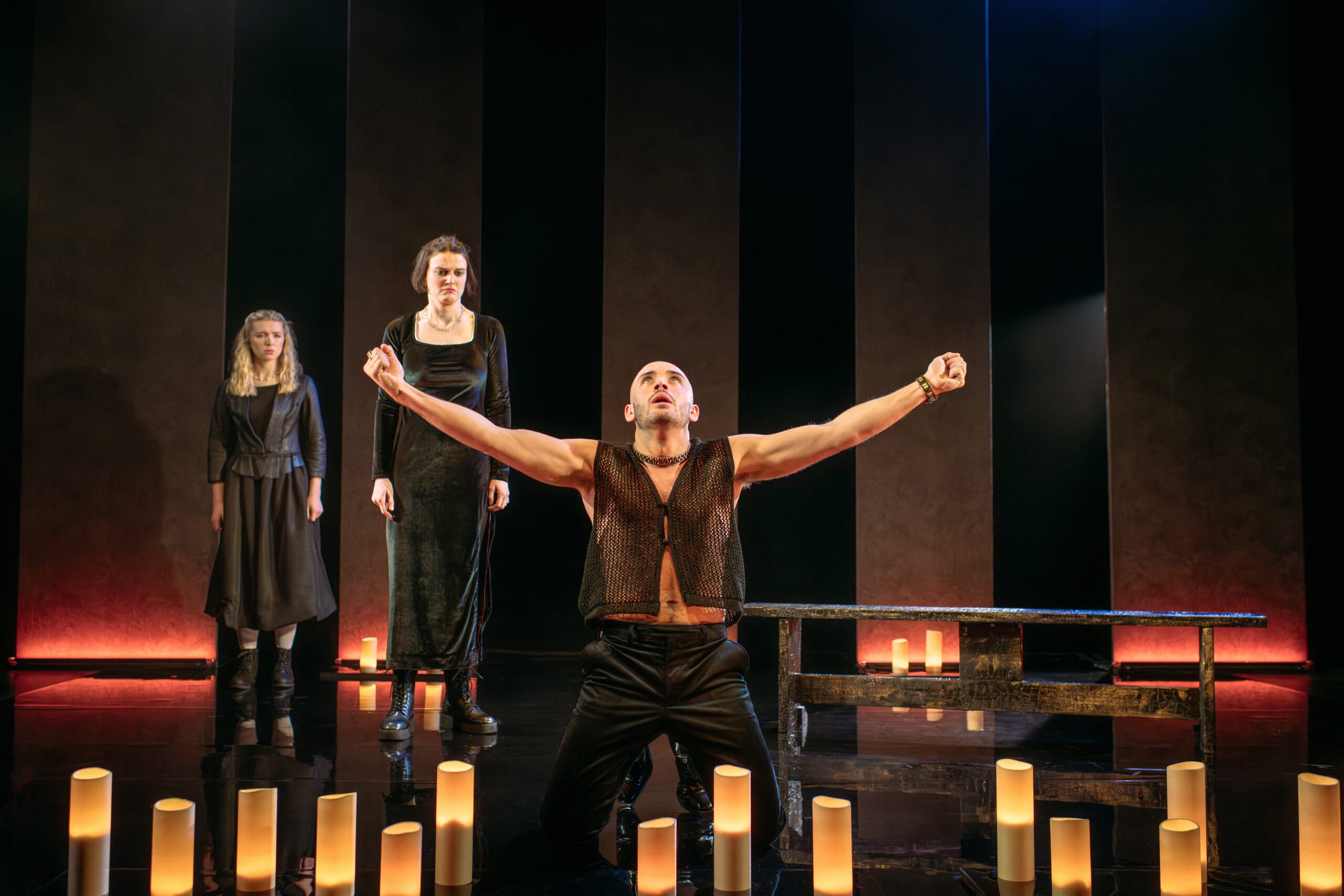 Three actors perform on stage; one man kneels with arms raised and two women stand behind him. The stage is dimly lit with cylindrical lights on the floor.