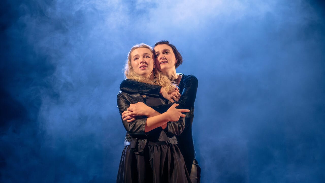 Two people stand closely together, with one person embracing the other from behind, both looking ahead with serious expressions against a smoky blue background.