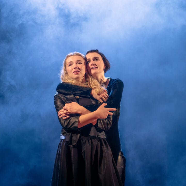 Two people stand closely together, with one person embracing the other from behind, both looking ahead with serious expressions against a smoky blue background.