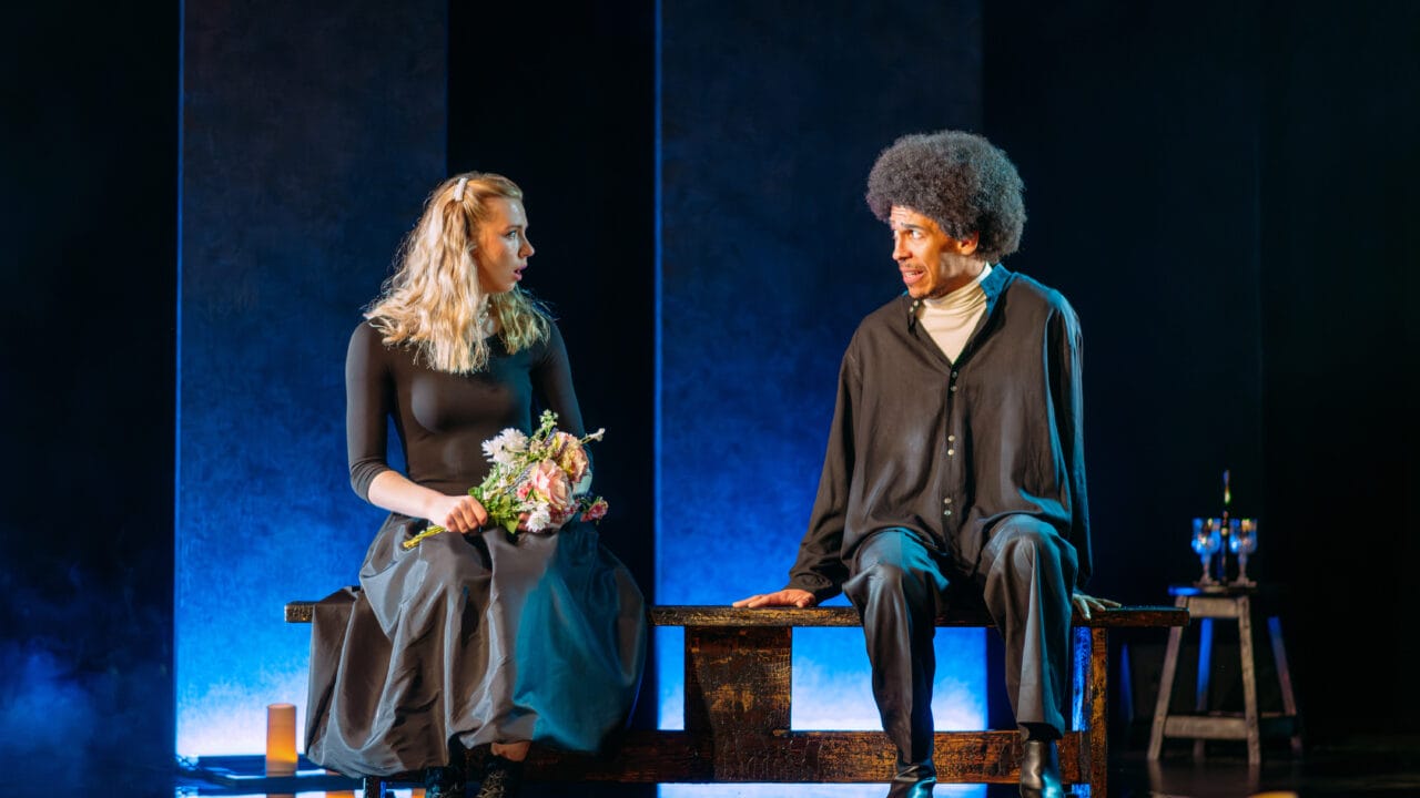 Two people sit on a bench onstage; the woman holds a bouquet of flowers, while the man looks at her. The lighting is dramatic, with candles on the floor and blue light in the background.