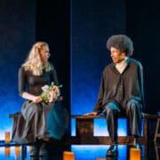 Two people sit on a bench onstage; the woman holds a bouquet of flowers, while the man looks at her. The lighting is dramatic, with candles on the floor and blue light in the background.