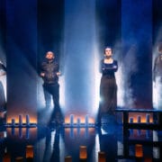 Four people stand on a dimly lit stage with dramatic lighting, surrounded by vertical light columns and candles, facing forward with serious expressions.