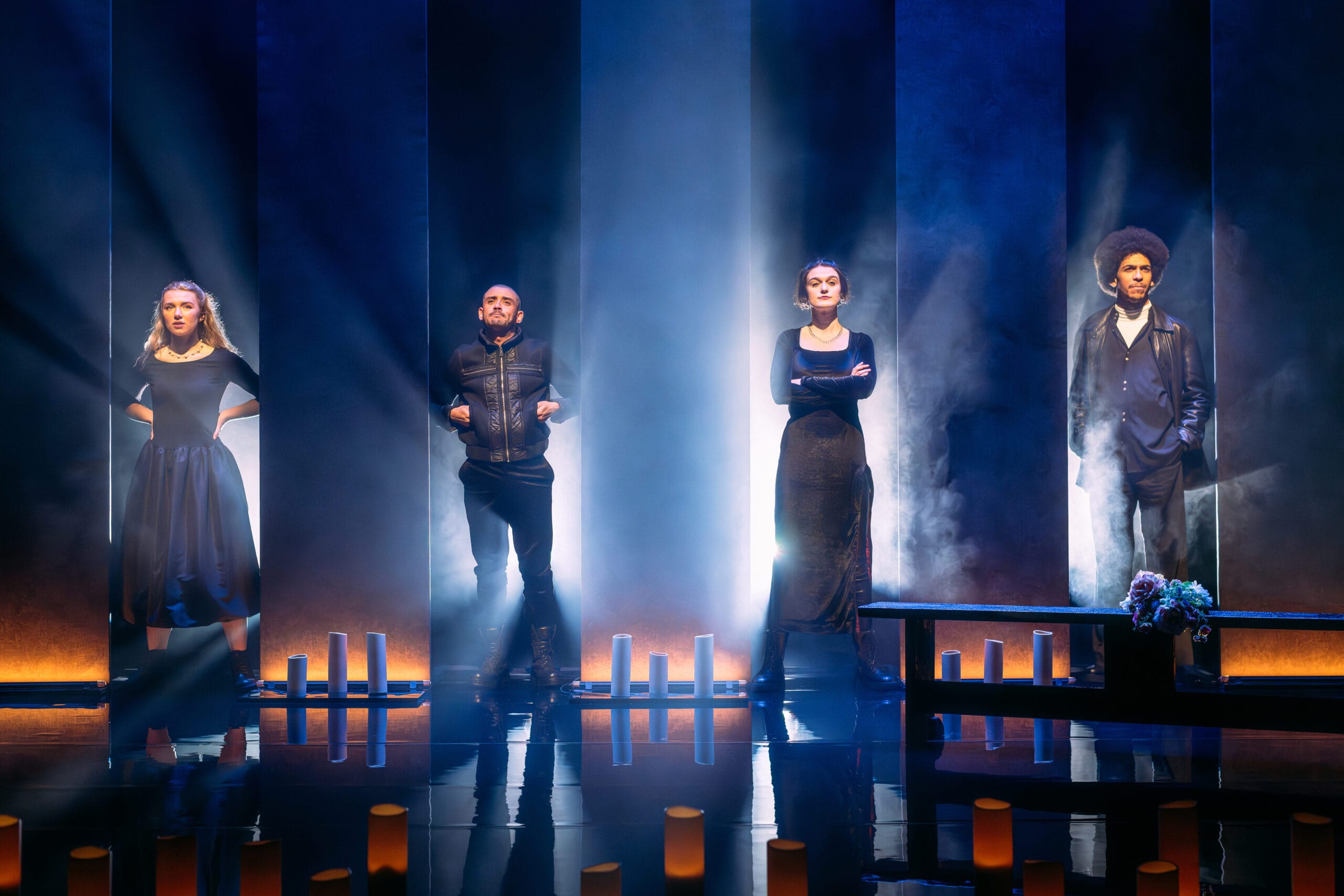 Four people stand on a dimly lit stage with dramatic lighting, surrounded by vertical light columns and candles, facing forward with serious expressions.