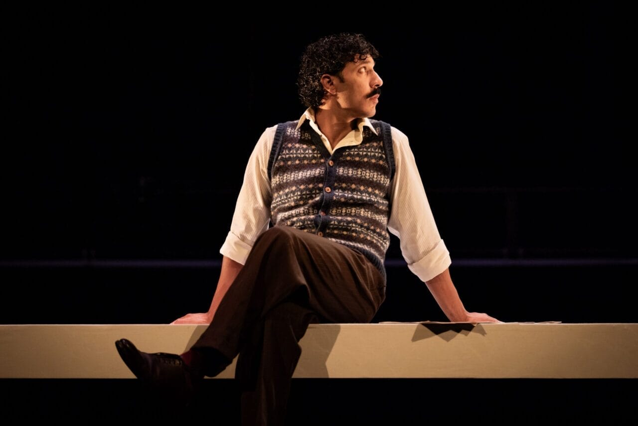 A person with curly hair, wearing a patterned vest, white shirt, and brown pants, sits on a ledge facing sideways in a dimly lit setting, evoking the mysterious atmosphere of a psychological thriller like The Talented Mr. Ripley.