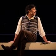 A person with curly hair, wearing a patterned vest, white shirt, and brown pants, sits on a ledge facing sideways in a dimly lit setting, evoking the mysterious atmosphere of a psychological thriller like The Talented Mr. Ripley.
