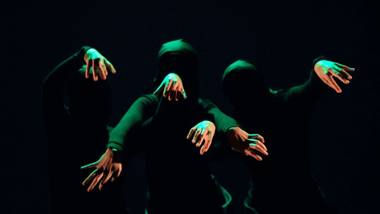 Three people in black clothing and masks extend their arms forward in dim green light against a dark background.