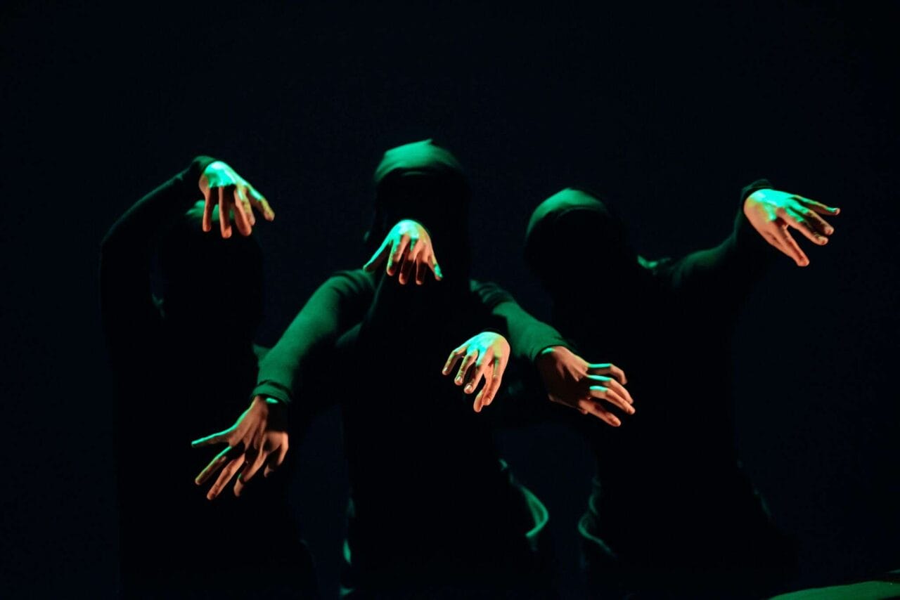 Three people in black clothing and masks extend their arms forward in dim green light against a dark background.