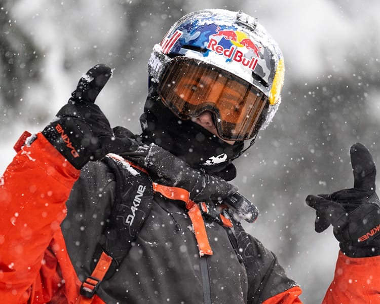 A person in orange winter gear and a Red Bull helmet poses in falling snow, making hand gestures with both hands. Snow covers their gloves and surroundings, embodying the adventurous spirit of the Blue Programme.