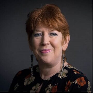 A woman with short red hair and long earrings is wearing a dark floral top, smiling in front of a dark background.
